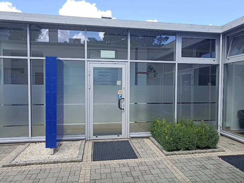 Przeszklone wejście do budynku z napisem „Laboratorium analityczne”, obok niebieska tablica z listą gabinetów lekarskich